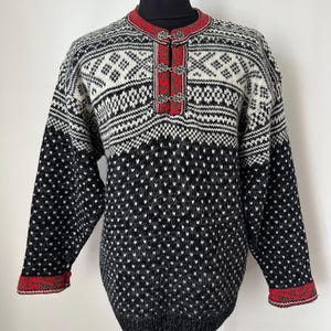 May include: A black, white, and red patterned sweater with long sleeves. The sweater features a round neckline with a red trim and decorative clasps. The cuffs and hemline have a red and green patterned trim.