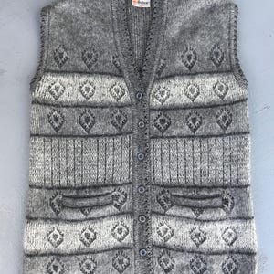 May include: A gray, button-down sweater vest with a diamond pattern and two front pockets. The vest has a V-neck and a label that reads "Osten". The knitted vest is a neutral color.
