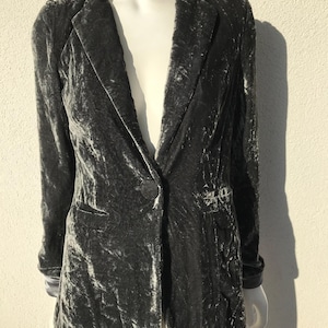 May include: A dark grey velvet blazer with a single-button fastening and notched lapels. The blazer features long sleeves and two front pockets. The fabric has a textured appearance.