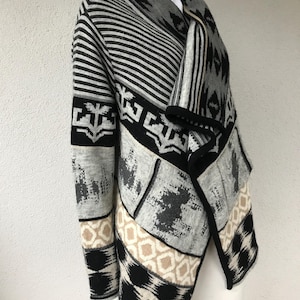Puede incluir: Un cárdigan con estampado gris, negro y blanco. El suéter presenta diseños geométricos, rayas y una parte delantera drapeada. Las mangas tienen una mezcla de patrones, incluyendo formas geométricas en blanco y negro y un patrón de diamantes beige y negro.