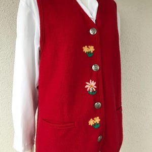 Puede incluir: Un chaleco de punto rojo con flores bordadas y botones plateados. El chaleco tiene un bolsillo en el lado izquierdo.