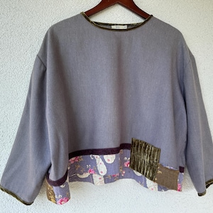 May include: A lavender long-sleeve top featuring a round neckline with olive green velvet trim. The hem includes a purple velvet band, a floral patterned fabric panel, and a rectangular olive green corduroy patch on the lower right side.