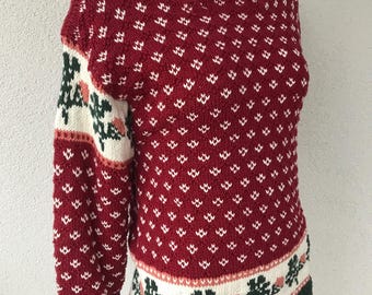 Könnte beinhalten: Ein roter Strickpullover mit weißem Rautenmuster und floralen Akzenten. Die Ärmel haben ein weißes Feld mit grünen und rosa floralen Designs. Der Saum und die Manschetten sind grün, und der Halsausschnitt ist gerippt rot.