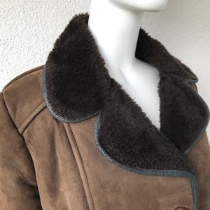 K&ouml;nnte beinhalten: Eine braune Wildlederjacke mit einem gro&szlig;en, dunkelbraunen Pelzkragen. Die Jacke hat einen doppelreihigen Verschluss und einen langen, taillierten Stil.