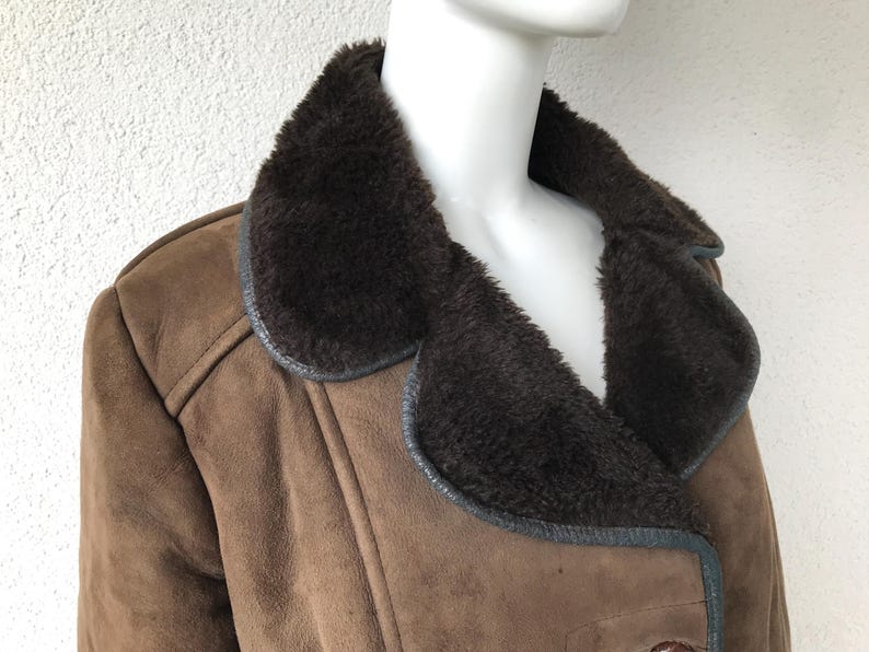 K&ouml;nnte beinhalten: Eine braune Wildlederjacke mit einem gro&szlig;en, dunkelbraunen Pelzkragen. Die Jacke hat einen doppelreihigen Verschluss und einen langen, taillierten Stil.