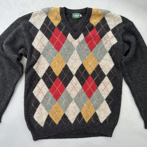 Peut inclure: Un pull à col en V et manches longues avec une base gris anthracite ornée d'un motif argyle multicolore composé de losanges rouges, jaune moutarde et gris clair. Le vêtement présente un col, des poignets et un ourlet côtelés, avec une étiquette verte CREW.