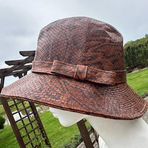 Puede incluir: Sombrero marrón y negro con estampado de piel de serpiente, ala ancha y lazo decorativo. El sombrero se exhibe en una cabeza de maniquí al aire libre, mostrando su textura y diseño. La paleta de colores del sombrero incluye tonos marrones y negros.