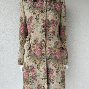 Op de afbeelding: Een jas met lange mouwen met een beige basis en een dicht bloemenpatroon in tinten roze, donkerrood, geel en olijfgroen. De jas heeft een gestructureerde kraag, een knoopsluiting aan de voorzijde met vijf donkere knopen en twee kleine klepzakken.