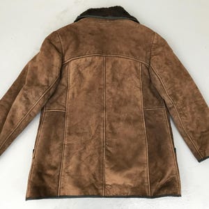 K&ouml;nnte beinhalten: Eine braune Wildlederjacke mit einem Lammfellkragen. Die Jacke hat einen Knopfverschluss und zwei Taschen.