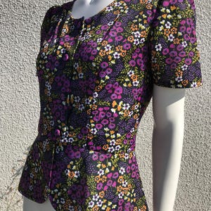 Può includere: Una blusa con stampa floreale viola, arancione, bianca e verde con chiusura a bottoni sul davanti e un orlo a peplo.