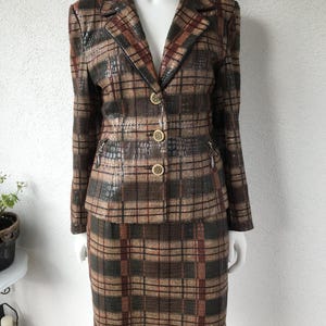 Könnte beinhalten: Ein zweiteiliger Vintage-Anzug mit einem karierten Muster in Braun-, Beige- und Burgundertönen. Die Jacke hat ein fallendes Revers, drei goldene Knöpfe und zwei kleine Reißverschlüsse. Der knielange Rock vervollständigt das klassische Ensemble.