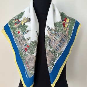 Puede incluir: Pañuelo cuadrado blanco con borde azul y ribete amarillo. El pañuelo presenta una ilustración detallada de un paisaje urbano con edificios, árboles y el texto "Diksmuide". El pañuelo está drapeado sobre un maniquí negro.