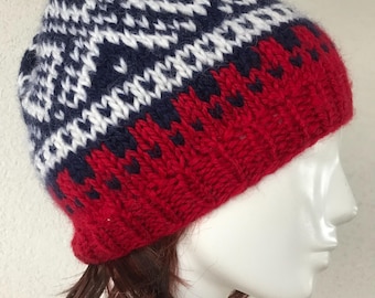May include: A knitted beanie hat with a geometric pattern. The hat features a red base, a navy blue band, and a white and navy blue patterned top. The hat is displayed on a mannequin head.