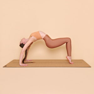 May include: A person performing a yoga pose called the upward bow or wheel pose on a brown cork yoga mat. The person is wearing a peach-colored sports bra and brown leggings.