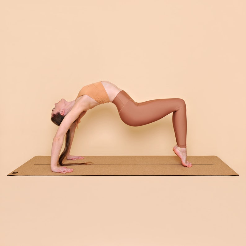 May include: A person performing a yoga pose called the upward bow or wheel pose on a brown cork yoga mat. The person is wearing a peach-colored sports bra and brown leggings.