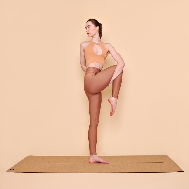 May include: A woman in a peach-colored sports bra and brown leggings is standing on a cork yoga mat. She is holding her right leg up and bending her knee, with her foot pointing towards her face.