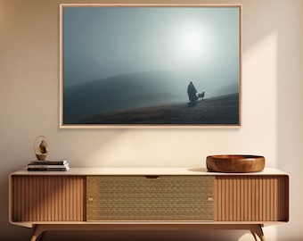 May include: A framed landscape print depicts a person and a dog on a hillside, bathed in a misty, atmospheric light. The artwork is set in a light-colored wooden frame, adding a touch of warmth to the scene. The print is displayed above a wooden cabinet.