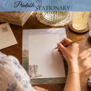 May include: A sheet of printable stationery featuring a landscape border with a pair of white cowboy boots decorated with colorful floral patterns at the bottom left corner. The paper is blank in the center, providing space for writing with a gold pen.