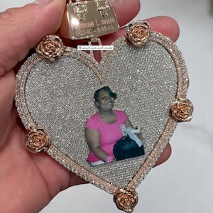 May include: A heart-shaped pendant with a photo of a woman in a pink shirt. The pendant is encrusted with small, sparkling stones and has rose-shaped accents. A small gold purse charm is at the top, with the name "Pashionee L. Moore" and dates engraved.