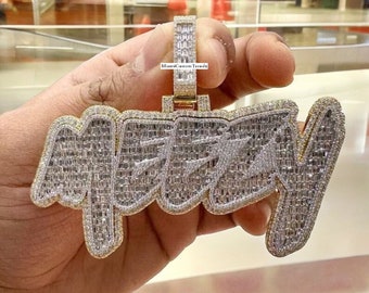 May include: A gold pendant with the word "Mezzy" spelled out in baguette-cut diamonds. The pendant is held in a hand and has a gold chain attached to it. The text "MiamiCustomTrends" is visible on the pendant.