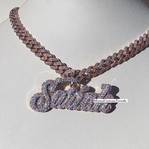 May include: A gold chain necklace with a diamond-encrusted pendant that says "Switz" in cursive. The chain is a Cuban link style.