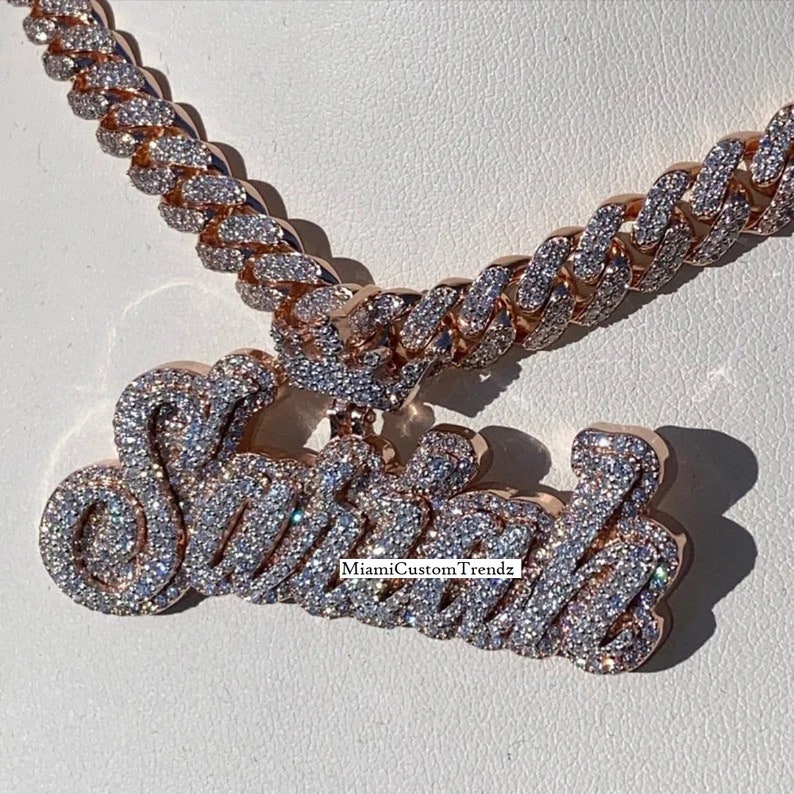 May include: A gold chain necklace with a large, custom-made pendant that says "Sasha" in sparkling diamonds. The chain is a Cuban link style and is also covered in diamonds.