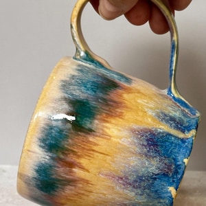 May include: A ceramic mug featuring a glossy glaze with a blended color palette of golden yellow, deep teal, and hints of purple. The mug has a smooth, rounded body and a curved handle, with the colors appearing to drip and swirl together.