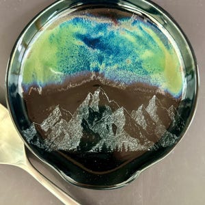 May include: A round, ceramic spoon rest with a mountain scene. The spoon rest features a dark blue rim and a glossy finish. The center has a mountain range design in white, with a blue and green sky. A silver spoon rests on the side.