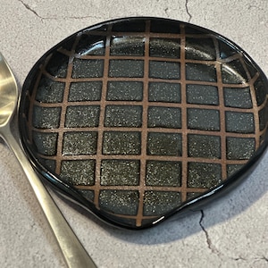 May include: A black ceramic spoon rest with a grid pattern in brown. The spoon rest has a slightly curved shape and a glossy finish. A silver spoon is partially visible on the left side of the image.