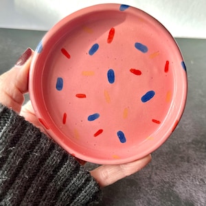 May include: A small, round, pink ceramic dish with a speckled design. The dish features blue, red, and yellow oblong shapes scattered across its surface. The dish is held in a hand, showcasing its size and design.