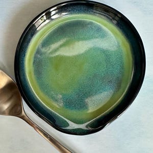 May include: A round ceramic spoon rest featuring a glossy glaze with a gradient of deep navy blue at the outer rim transitioning into a vibrant lime green and teal center. A polished stainless steel spoon rests partially beside the dish.