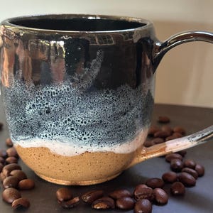 May include: A ceramic mug with a glossy black upper section that transitions into a textured, bubbly white glaze, ending in a speckled tan base. The mug features a curved handle and is surrounded by scattered roasted coffee beans.