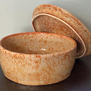 May include: A round ceramic container with a matching lid, featuring a glossy beige glaze with a speckled orange and brown pattern. The container has a low profile and straight sides, while the lid is tilted against the back rim.
