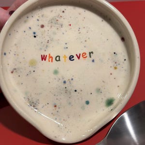 May include: A white ceramic spoon rest with a speckled glaze and the word "whatever" written in rainbow colors.