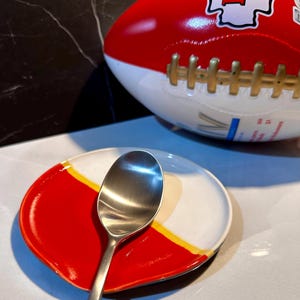 May include: A red and white ceramic spoon rest with a gold stripe. The spoon rest is shaped like a half-circle and has a spoon resting on it.