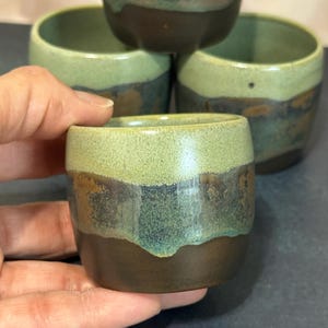 May include: A set of four small ceramic cups featuring a two-tone glaze. The upper portion of each cup is a light sage green, while the lower portion is a dark brown with mottled blue and green accents where the two colors meet and overlap.