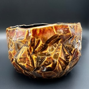 May include: A brown and gold ceramic bowl with a textured, craggy surface. The bowl has a wide, rounded shape and a slightly uneven rim.