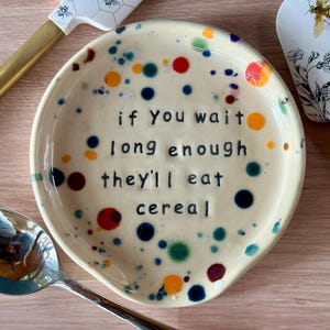 May include: A round, cream-colored ceramic spoon rest featuring a colorful pattern of small, multi-colored dots. The center of the dish is stamped with the black text: if You wait long enough they'll eat cereal. A silver spoon is positioned nearby.
