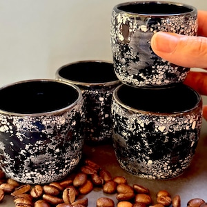 May include: A set of three small ceramic cups featuring a black interior and a textured exterior with a mottled black and white speckled pattern. The cups are cylindrical with a smooth rim and are surrounded by scattered roasted coffee beans.