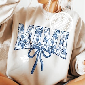 Pode incluir: Camisola creme com a palavra "MAMA" em estampa floral azul. Uma fita azul está abaixo da palavra. O logotipo The Crafty Shop está na parte inferior esquerda. O selo Original está no canto superior direito.