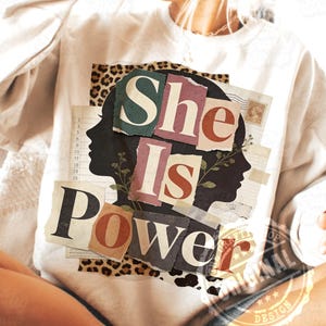 Può includere: Una felpa bianca con la scritta "She Is Power" in stile collage. Il design include la silhouette di un profilo femminile, elementi floreali e un bordo con stampa leopardata. La felpa è uno stile casual e confortevole.