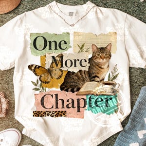May include: White t-shirt with the text "One More Chapter" and a graphic of a cat, butterfly, and open book. The design includes a coffee cup and botanical elements. The shirt has a crew neck and short sleeves.