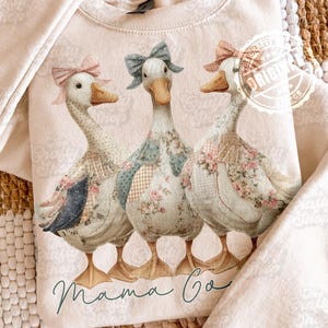 Pode incluir: Camisola bege com uma ilustração extravagante de três gansos adornados com tecidos florais e estampados, cada um com um laço. O texto "Mama Co" está escrito em itálico abaixo dos gansos. A camisola tem gola redonda e mangas compridas.