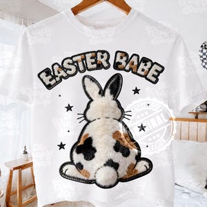 Może przedstawiać: Biała koszulka z napisem "EASTER BABE" nad wzorem królika. Królik ma puszysty, wielokolorowy wzór futra i jest otoczony czarnymi gwiazdami. Koszulka wisi na drewnianym wieszaku.