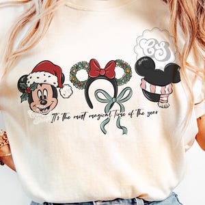 May include: Cream-colored t-shirt featuring three Mickey Mouse-themed holiday designs. The designs include Mickey with a Santa hat, a wreath headband with a red bow, and Mickey with a pink and white scarf. The text "It's the most magical Time of the year" is also printed on the shirt.