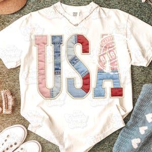 Pode incluir: Camiseta branca com a palavra "USA" em letras de patchwork. As letras são feitas de tecido estilo jeans vermelho, branco e azul. A letra "A" apresenta um design de selo com a Estátua da Liberdade. Um colar de prata é visível.
