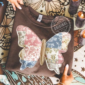 May include: A brown sweatshirt featuring a large butterfly applique. The butterfly wings are made of patchwork fabric in shades of pink, blue, and cream with floral patterns. The sweatshirt has the text "The Crafty Shop Original".