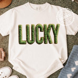 Pode incluir: Camiseta branca com a palavra "LUCKY" em letras verdes grandes e texturizadas. A palavra é delineada em dourado. A camiseta tem gola redonda e mangas curtas. A imagem também inclui um chapéu marrom, tênis brancos e uma meia azul.