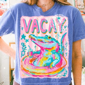 Può includere: T-shirt blu a maniche corte con una grafica colorata. La grafica presenta la parola "VACAY" in rosa, un alligatore dei cartoni animati in un salvagente rosa e giallo e il testo "The Gritty Shop".