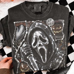 May include: A charcoal gray short-sleeve t-shirt featuring a graphic print of a hooded figure with a mask holding a knife, surrounded by smaller images of pumpkins and the word Halloween. The shirt has a Comfort Colors brand label at the neck.